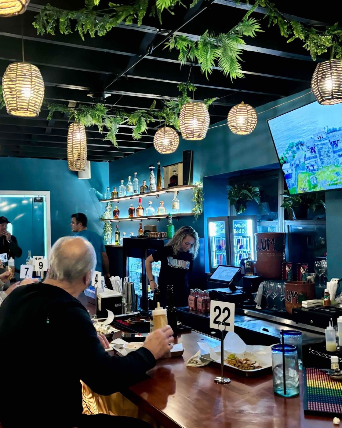 Tulum staff member behind the bar, tequila shelf lit warmly with hanging greenery overhead