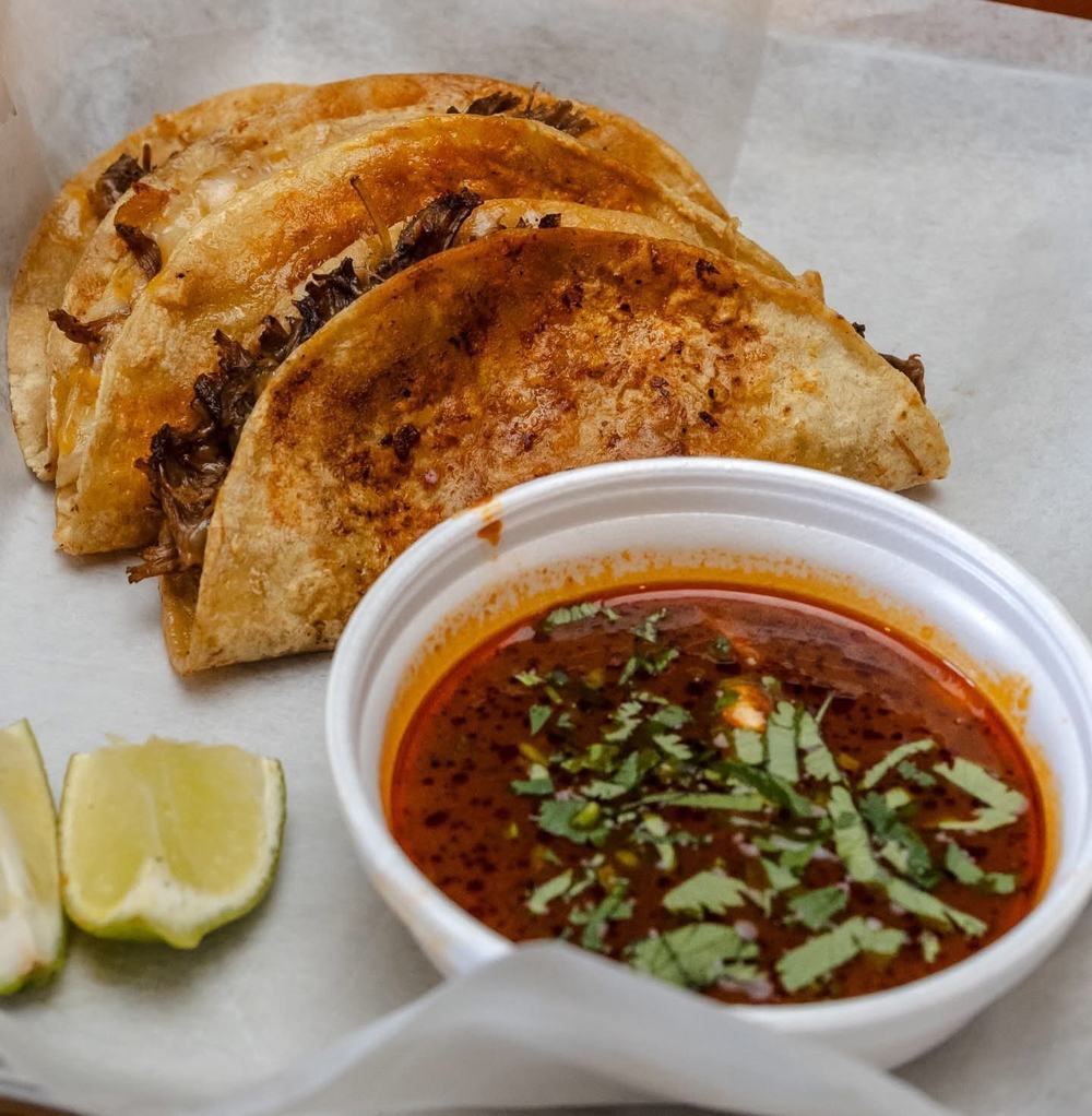 Birria tacos with melted cheese, served with a cup of consommé for dipping and lime wedges
